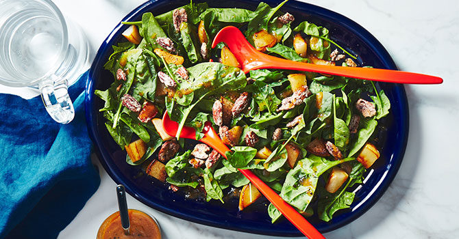 spinach salad topped with sliced pears and candied nuts in a blue bowl with orange tongs and a side of dressing