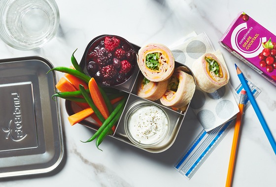 Bento lunch box on marble background. Filled with ham and cheese wrap, veggies and dip, plus a field berry compote.