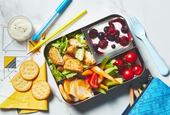 Bento lunch box on marble background. Filled with kids lunchbox staples, including crispy chicken salad, cheese and crackers and field berry yogurt.