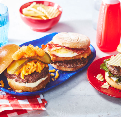 Table with three burgers – triple mac and cheese burger, chips n dip burger, and the brekkie burger – on plates, with a drink, bottle of ketchup and bowl of chips in the background.
