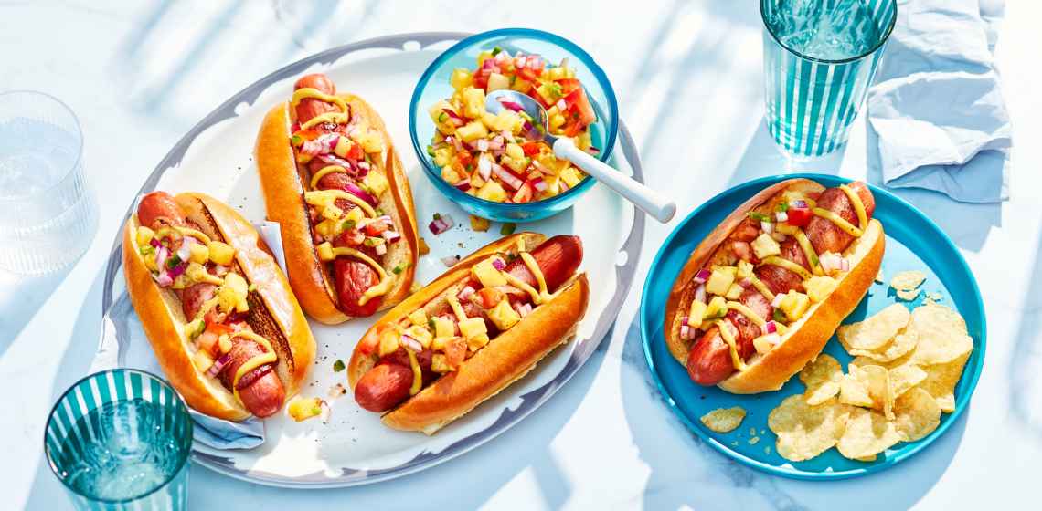 blue platter of three loaded hot dogs with one side plate of a hot dog and chips to the side