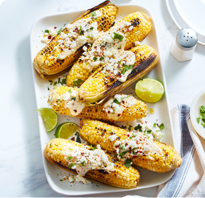 White table with white serving platter of grilled corn with dressing and herbs on top