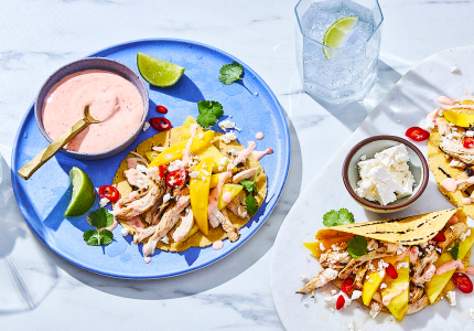 Serving board with tropical chicken and mango tacos, plus blue dinner plate to the side with chicken taco and bowl of chili-lime crema.