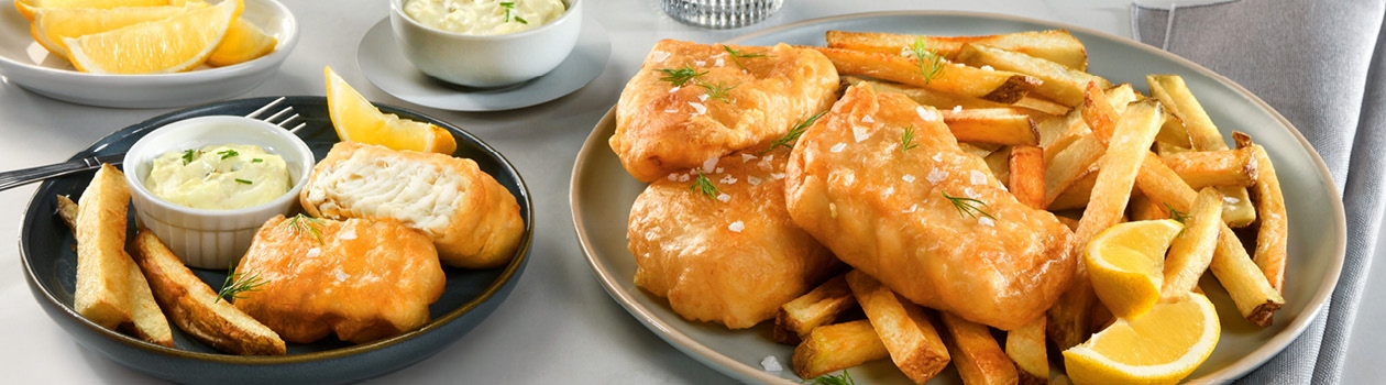 A big plate of fish and chips with a couple slices of lemon and a smaller plate with a fried piece of fish broken open, chips, a fork, a slice of lemon and some tartar sauce.
