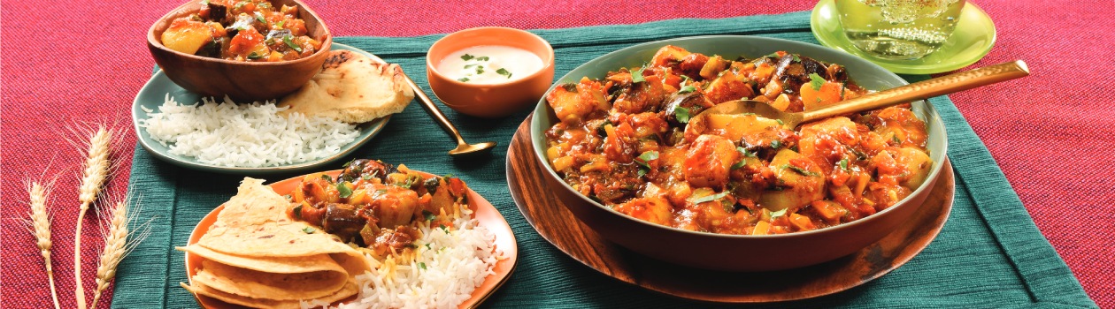 A big bowl of curry with a spoon, two smaller plates with curry, rice, and naan/roti, a small bowl of tadka, and wheat stalks for decoration on the side.